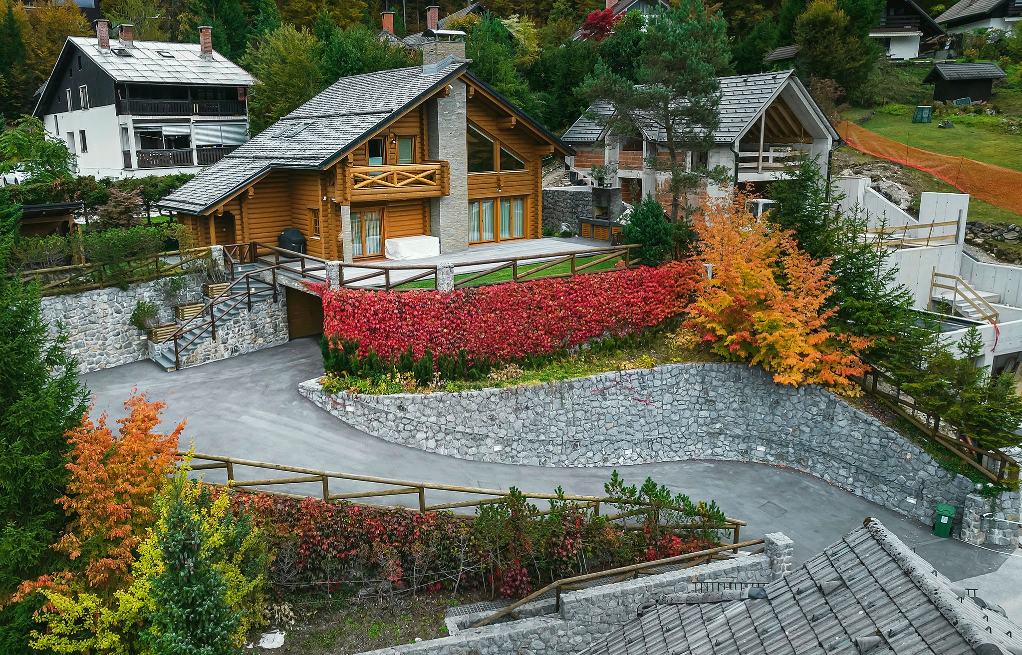 Elevated three-quarter aerial view of the chalet, with crimson autumn creeper on the stone retaining wall and the driveway curving toward the house.