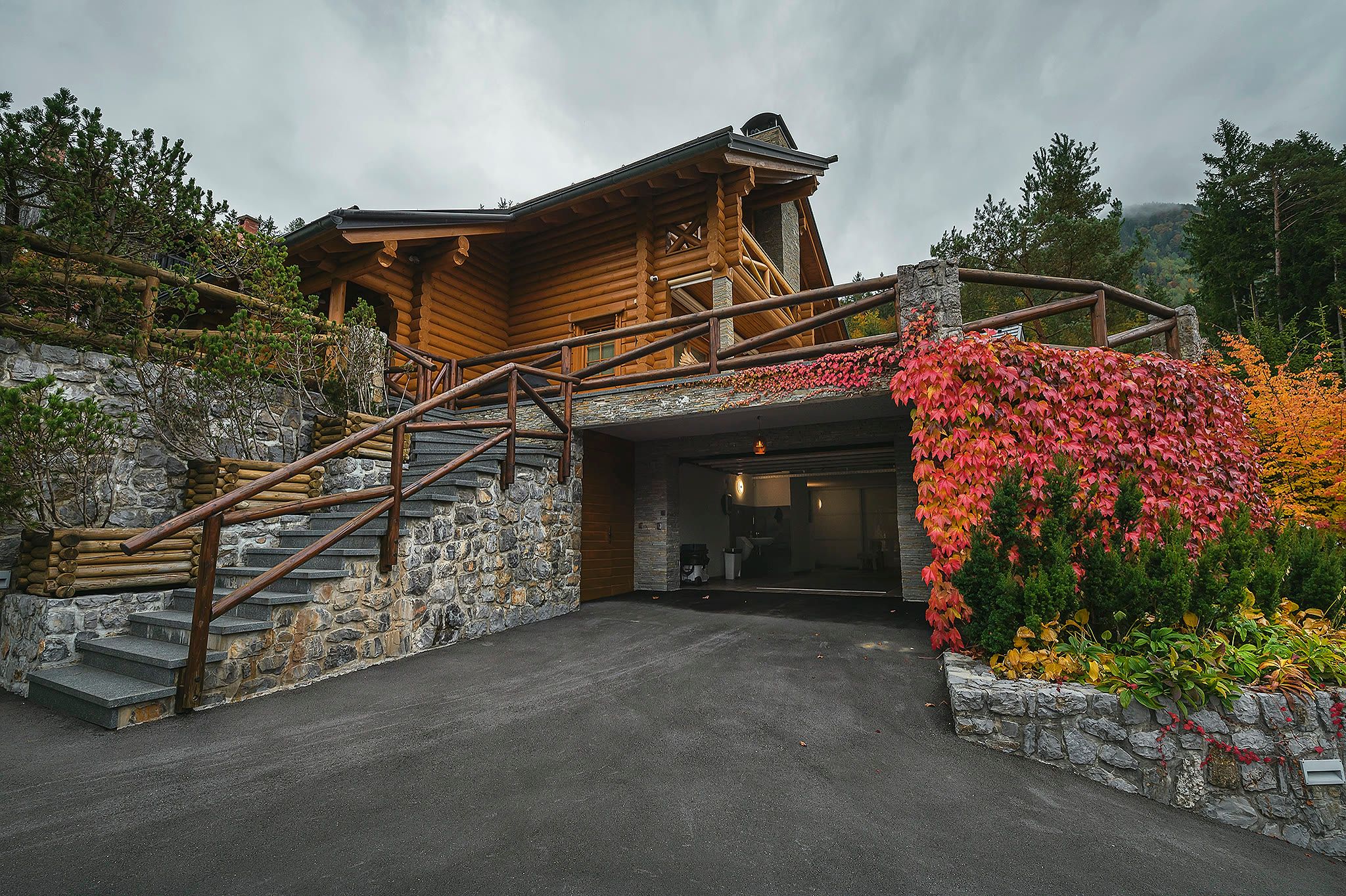 Ground-level exterior view of the chalet with the garage entrance, a wide concrete driveway, a timber stair, and autumn creeper on the wall.