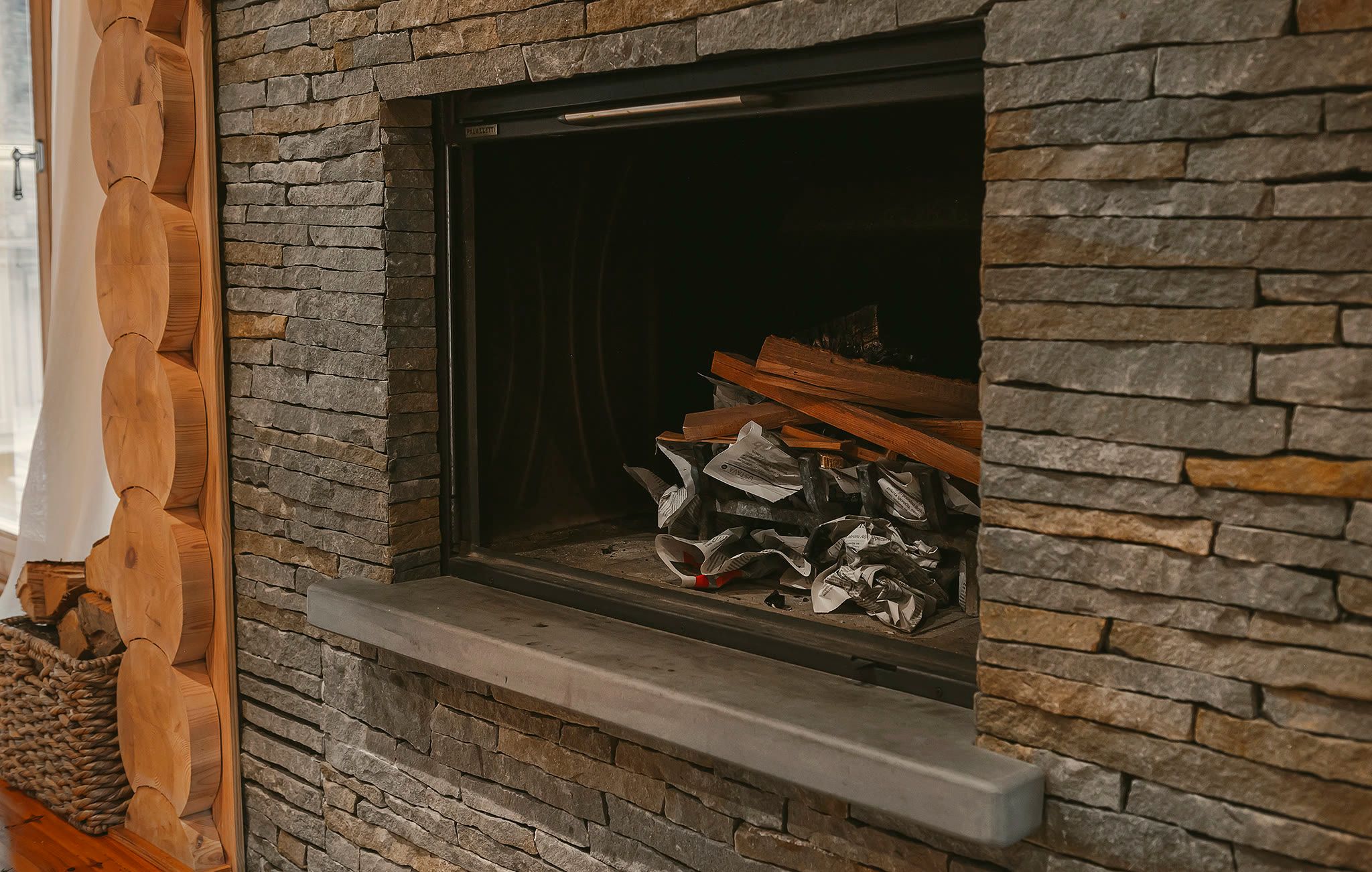 Detail of the stone fireplace, showing the firebox with stacked logs, a dry-stone surround, and a concrete lintel.