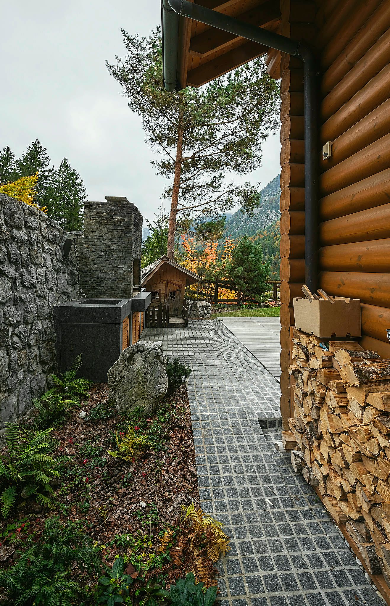 A side path with firewood stacked against the round-log wall next to a stone wall.