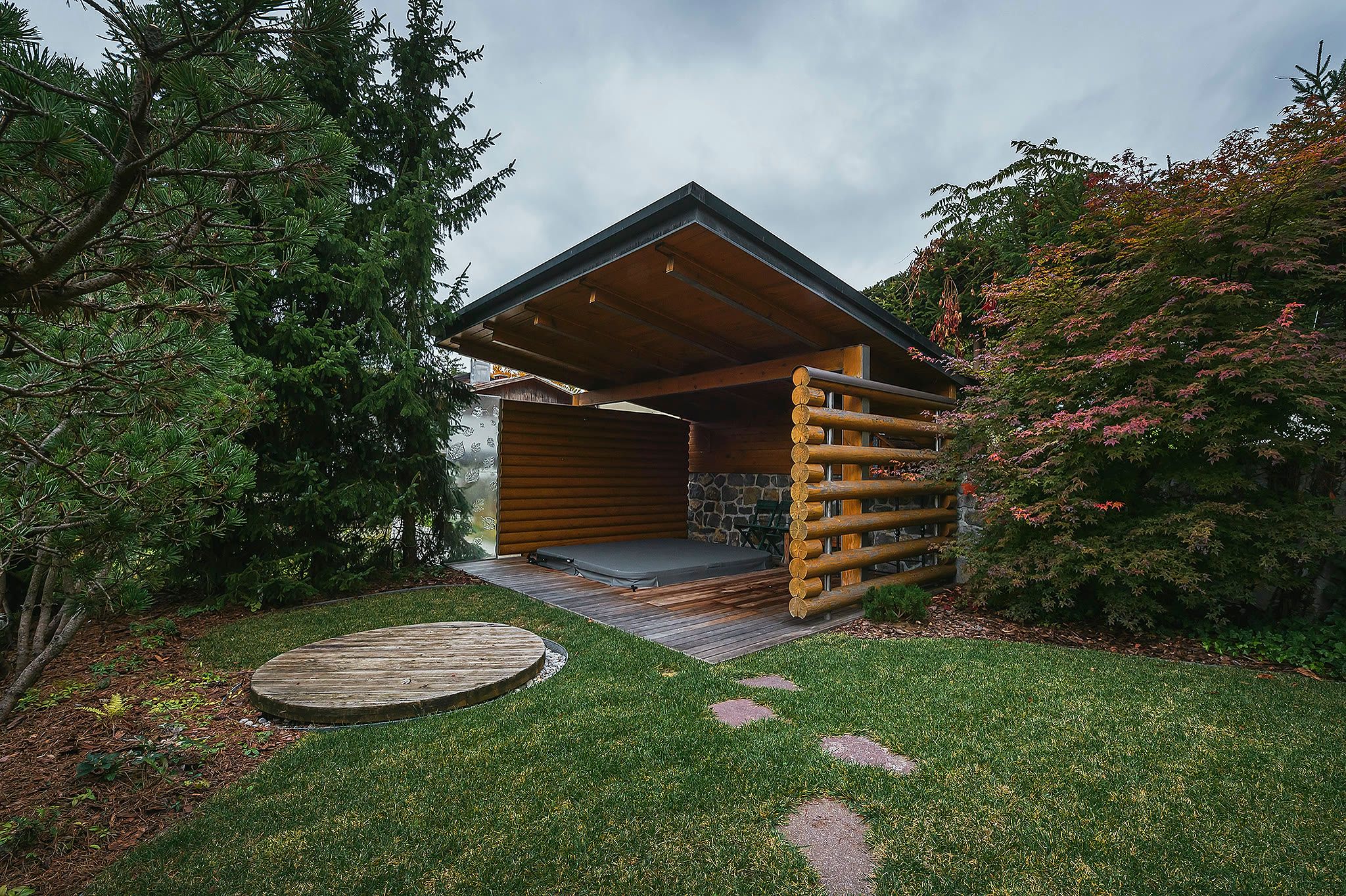 The garden with a lawn, stepping stones, a covered circular hot tub, and a timber-clad outdoor structure.