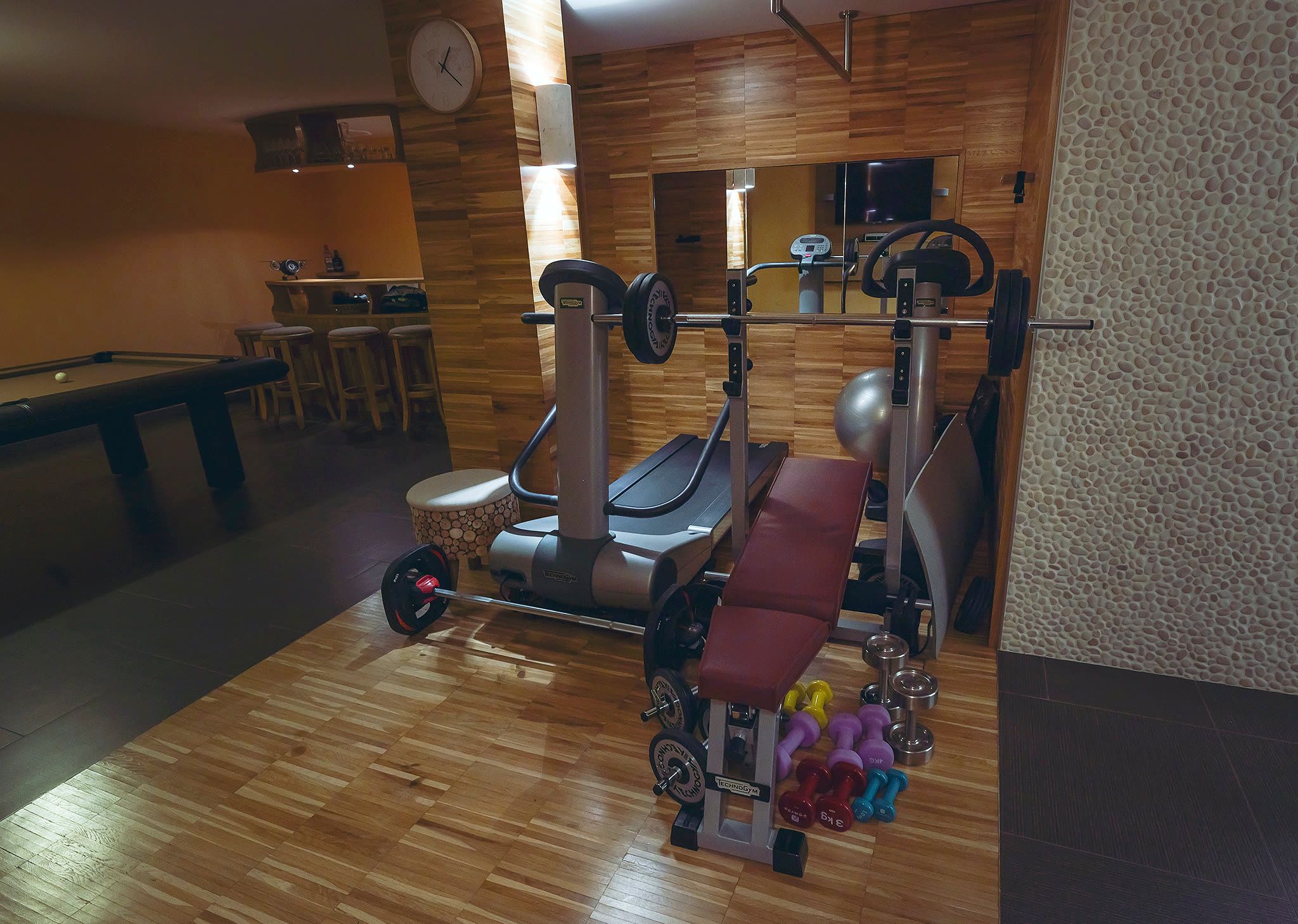 The gym with a cross-trainer, a weight bench, dumbbells, a timber floor, and a bar behind.