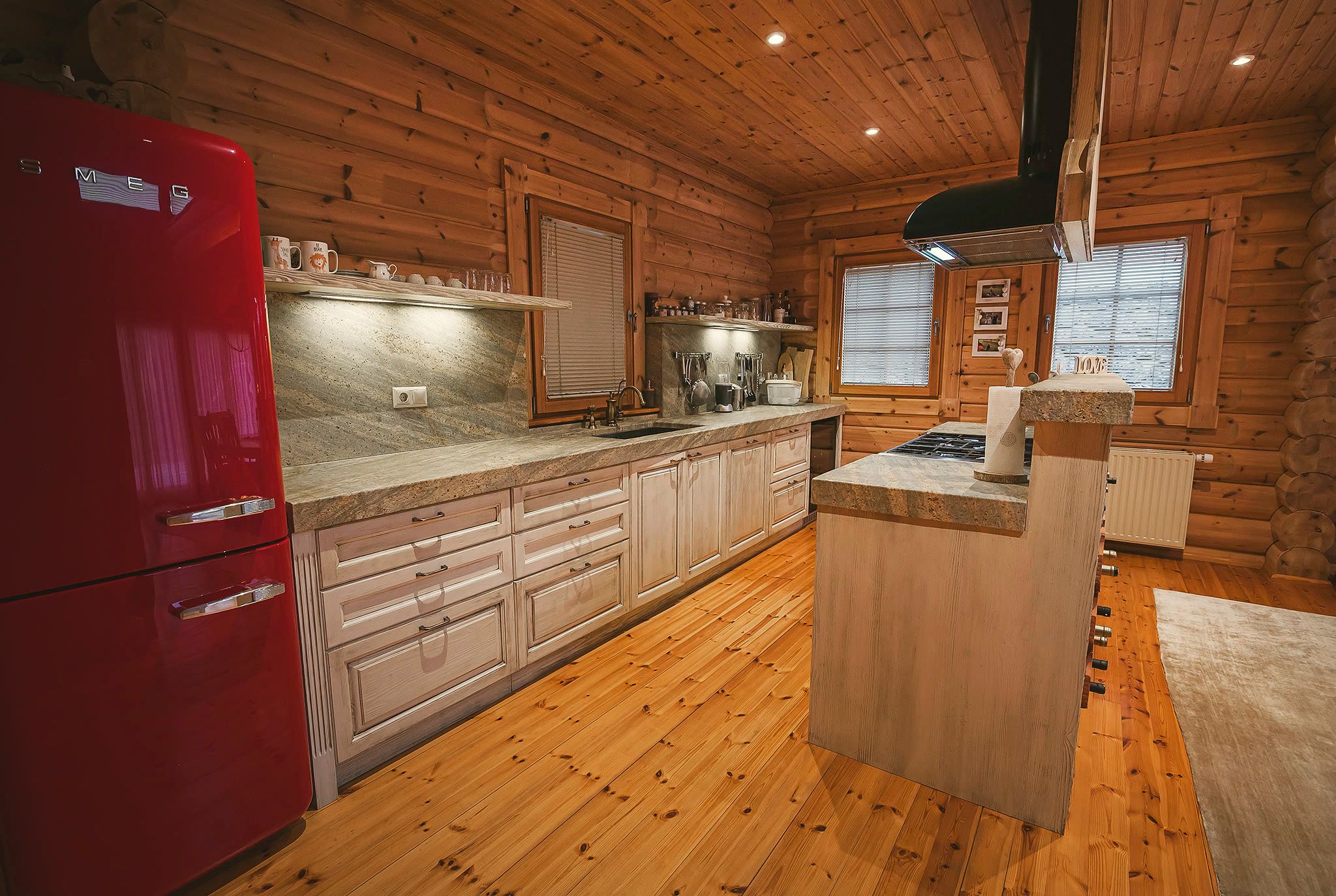 The kitchen with cream cabinetry, a stone worktop, a red retro fridge, and a timber floor around the island.