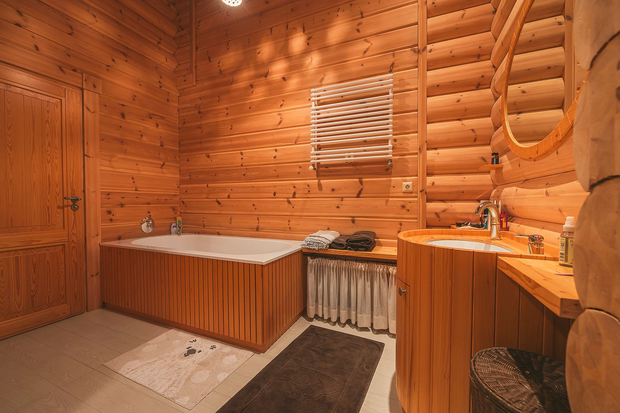 A log-walled bathroom with a timber-panelled bath, a basin, a round mirror, and a heated towel rail.