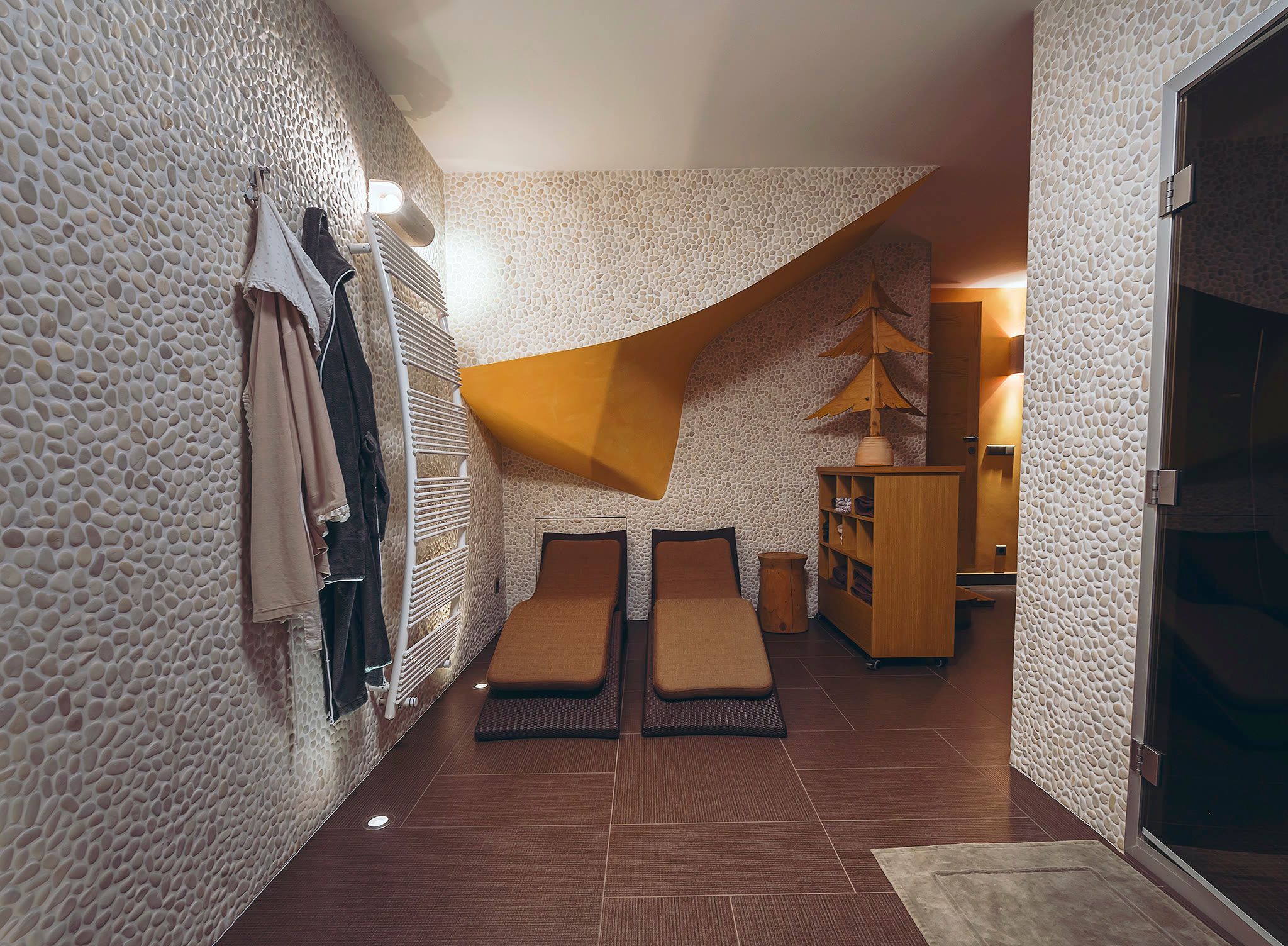 The spa relaxation area with two loungers, robes on hooks, a pebble-textured wall, and a curved timber screen.
