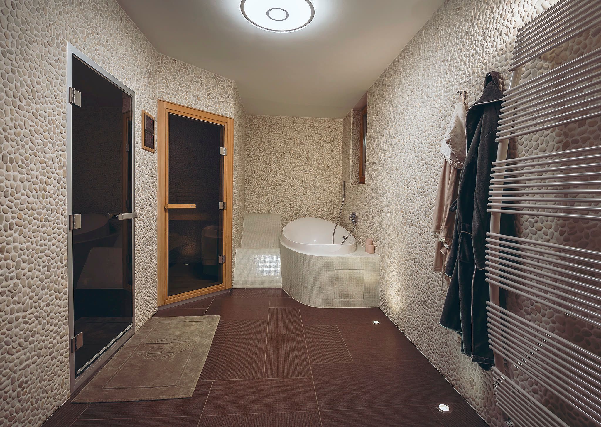 The spa area with a freestanding soaking tub, robes, a heated towel rail, a dark tiled floor, and sauna doors beyond.